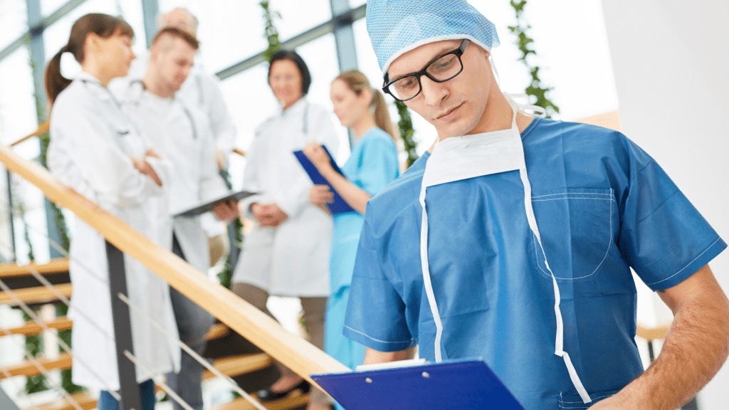 A man in scrubs stands confidently, holding a clipboard, ready to take notes in a medical setting.